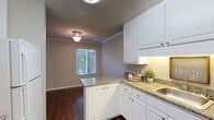 This single-story residence offers two bedrooms and one bathroom across a thoughtfully designed floor plan. The kitchen is a standout feature, showcasing crisp white cabinetry with brushed nickel hardware, granite-look countertops, and under-cabinet lighting that adds warmth and practicality to the workspace. A stainless steel sink and ample upper and lower cabinet storage make this kitchen both functional and visually appealing. The kitchen flows naturally into an adjacent dining area, defined by rich dark hardwood-style flooring that extends throughout the living spaces, creating a cohesive and inviting atmosphere. A window in the dining area allows natural light to filter in through slatted blinds, brightening the space while maintaining privacy. Crown molding accents the transition between walls and ceiling, adding a refined touch to the interior. Textured ceilings and flush-mount light fixtures provide a classic, understated aesthetic. The neutral wall tones throughout the space complement the white cabinetry and dark flooring, creating a balanced and versatile palette suitable for a variety of décor styles.