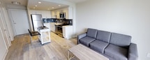 This well-designed single-floor space presents a seamless open-concept layout that connects the kitchen and living area, creating a cohesive and functional environment. The kitchen is outfitted with stainless steel appliances, including a refrigerator, range, and microwave, complemented by crisp white cabinetry and a stylish dark tile backsplash that adds a modern contrast. A stainless steel sink sits beneath recessed lighting, and the counter space offers ample room for meal preparation. A portable kitchen cart provides additional storage and flexibility. The living area flows naturally from the kitchen, anchored by a contemporary gray sofa and a minimalist wood-and-metal coffee table, creating a relaxed and inviting atmosphere. Light wood-toned flooring runs throughout the space, lending warmth and visual continuity to the overall design. The clean white walls and recessed ceiling lights contribute to a bright and airy ambiance. The overall aesthetic is modern and understated, with a neutral palette that offers a versatile backdrop. The thoughtful layout maximizes the available space, making it both practical and comfortable for everyday living.