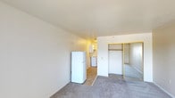 Unit #246 at 2525 Highland Avenue in National City is a single-floor studio offering a straightforward and functional layout ideal for efficient living. The main living area features wall-to-wall carpet flooring and is anchored by a mirrored sliding-door closet with a gold-tone frame, providing generous storage and a sense of added depth to the space. A full-size refrigerator sits conveniently at the transition between the living area and the kitchenette, which is visible in the background and includes a range and cabinetry for everyday cooking needs. The walls throughout are finished in a clean, neutral white, complemented by a textured ceiling that adds subtle character. The open-concept design allows the living and kitchen areas to flow naturally into one another, making the space feel cohesive and easy to navigate. Overall, the unit presents a clean, no-frills interior well-suited for a single occupant seeking a practical and low-maintenance residence in National City.