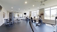 Located at 700 Citation Lane in Cherry Hill, New Jersey, this fitness center offers a clean, modern environment designed to support a full range of workout routines. The space is outfitted with an impressive selection of cardio equipment, including elliptical machines, stationary bikes, and treadmills, all arranged to maximize floor space and ease of movement. Strength training options are also available, with weight machines and free weight stations positioned along the mirrored wall, which enhances the sense of openness throughout the room. Large windows line the perimeter, flooding the interior with abundant natural light and providing views of the surrounding greenery outside. The dark, rubberized flooring contrasts cleanly with the bright white ceiling, which features recessed lighting and ceiling fans to ensure consistent air circulation and a comfortable workout environment. A wall-mounted flat-screen television is conveniently positioned for viewing during exercise. The overall ambiance is bright, functional, and well-maintained, reflecting a thoughtfully designed amenity space suited for residents seeking a convenient and fully equipped place to stay active.