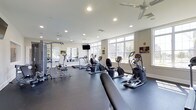 Located at 700 Citation Lane in Cherry Hill, New Jersey, this fitness center offers a clean, modern environment designed to support a full range of workout routines. The space is outfitted with an impressive selection of cardio equipment, including elliptical machines, stationary bikes, and treadmills, all arranged to maximize floor space and ease of movement. Strength training options are also available, with weight machines and free weight stations positioned along the mirrored wall, which enhances the sense of openness throughout the room. Large windows line the perimeter, flooding the interior with abundant natural light and providing views of the surrounding greenery outside. The dark, rubberized flooring contrasts cleanly with the bright white ceiling, which features recessed lighting and ceiling fans to ensure consistent air circulation and a comfortable workout environment. A wall-mounted flat-screen television is conveniently positioned for viewing during exercise. The overall ambiance is bright, functional, and well-maintained, reflecting a thoughtfully designed amenity space suited for residents seeking a convenient and fully equipped place to stay active.