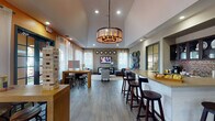 This single-floor community lounge space is thoughtfully designed to encourage social interaction and relaxation. The open-plan layout flows seamlessly between distinct activity zones, all unified by wood-look tile flooring and a cohesive warm color palette accented by bold orange walls.

A central gathering area anchors the space beneath a dramatic vaulted ceiling with a statement drum chandelier. This zone features a wall-mounted flat-screen TV flanked by sheer white curtains and large windows that fill the room with natural light. A gray sectional sofa, leather accent chairs, a geometric-patterned area rug, and a dark wood coffee table complete the lounge seating.

Adjacent to the lounge is a dedicated game area equipped with oversized tabletop games, including a giant Jenga tower and Connect Four, arranged around bar-height seating. A built-in bar counter with bar stools provides an additional social hub, while a fully equipped kitchenette with dark cabinetry, stone-look countertops, a mosaic tile backsplash, and glass-front upper cabinets supports food and beverage service.

Throughout, recessed lighting supplements the abundant natural light from multiple windows, creating a bright and welcoming ambiance suited for both casual gatherings and organized community events.