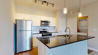 Located at 291 Graceland Blvd in Columbus, Ohio, this well-designed one-bedroom, one-bathroom residence offers a thoughtfully organized single-floor layout that maximizes functionality and comfort. The open-concept living area seamlessly connects the living room, dining space, and kitchen, creating a cohesive and versatile environment. The kitchen is a standout feature, equipped with stainless steel appliances including a refrigerator, range, and microwave, complemented by crisp white cabinetry, a subway tile backsplash, and a central island with dark granite countertops and a built-in sink — all illuminated by pendant and track lighting overhead. Hardwood-style flooring runs throughout the kitchen and bathroom, while plush carpet covers the living and bedroom areas. The bedroom features natural light from a large window and a sliding glass door leading to a private balcony with outdoor views. The bathroom offers a wood-toned floor, a walk-in shower, a dark granite vanity with white cabinetry, and direct access to a spacious walk-in closet with wire shelving. Additional highlights include three closets for ample storage and an in-unit laundry area, rounding out this efficiently planned and modern residence.
