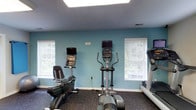 This single-level fitness room offers a focused and functional workout environment designed to accommodate a variety of cardio-based exercise routines. The space is finished with teal-painted walls that lend a fresh, energizing atmosphere, complemented by dark rubber flooring that provides durability and traction underfoot. Overhead, large recessed panel lighting ensures the room is well-lit throughout the day.

Natural light filters in through multiple white-framed windows fitted with horizontal blinds, offering views of surrounding greenery while allowing users to control light levels as needed. The windows are evenly distributed along the walls, contributing to an open and airy feel within the space.

The room is equipped with a selection of modern cardio machines, including a recumbent bike, an elliptical trainer, and a treadmill, each featuring an integrated display monitor for tracking workout metrics. The equipment is thoughtfully arranged to allow comfortable spacing between machines, ensuring ease of movement and an unobstructed workout experience. A small informational sign is mounted on the wall, adding a practical touch to the overall layout.