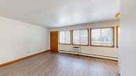 This efficiently designed single-floor studio offers a practical and well-organized layout comprising a combined kitchen and living area, a closet, and a full bathroom. The open living space is finished with durable vinyl plank flooring in a warm wood tone, white walls, and classic wood-trimmed baseboards and door frames that add a consistent decorative detail throughout. Multiple windows fitted with horizontal blinds line the living area, allowing ample natural light to fill the room while offering views of the surrounding outdoor greenery. A wall-mounted air conditioning unit and baseboard heating system ensure year-round climate comfort. The kitchen is equipped with warm-toned wood cabinetry extending to the ceiling for generous storage, a white electric range, a stainless sink with a functional countertop workspace, and a full-size refrigerator. The bathroom features a full bathtub and shower combination with clean white surround panels and warm wood trim framing. A closet provides dedicated storage space. The unit's entry door opens directly into the main living area, with a clear sightline through to the bathroom and closet beyond, making the layout easy to navigate. Overall, the space presents a straightforward, functional design with consistent finishes and practical amenities throughout.