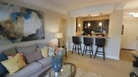 This single-floor residence showcases an open-concept design that seamlessly connects the living area to the kitchen, creating a cohesive and functional living space. The living room features neutral-toned carpet, crown molding, and warm beige walls that lend a clean, polished aesthetic. A comfortable gray sofa anchored by a glass-top coffee table and a textured area rug anchors the seating area, complemented by colorful accent pillows that add visual interest. A large abstract artwork adorns one wall, contributing an artistic focal point to the space.

The kitchen is accessible via a raised bar counter finished with granite-style countertops, lined with three sleek black swivel bar stools that offer casual dining and entertaining options. Dark cabinetry provides a bold contrast against the lighter walls, while three pendant lights hang above the bar, casting warm, focused illumination. Black appliances complete the kitchen's contemporary look. Crown molding details continue into the kitchen area, reinforcing the consistent, refined character of the space. Overall, the residence strikes a balance between comfort and sophistication.