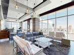 Located in the heart of Charlotte's Brooklyn Village neighborhood, this upscale amenity lounge delivers a striking blend of contemporary design and urban sophistication. The interior is anchored by dramatic floor-to-ceiling windows that flood the space with natural light while framing sweeping views of the Charlotte skyline. The open layout features a large, elegantly appointed bar with dark stone countertops, textured tile cladding, and a row of sleek white barstools, complemented by pendant lighting throughout. Distinct lounge seating areas are defined by bold geometric rugs and a mix of leather sofas, chrome-accented chairs, and glass coffee tables, creating an inviting yet modern atmosphere. Deep burgundy accent walls and layered wood-paneled columns add warmth and visual depth. A separate transitional lounge near the entry showcases soaring ceilings and bold black furnishings. The space extends outdoors to a generous rooftop terrace, where glass railings, manicured hedges in planters, and ample white lounge seating take full advantage of the open-sky setting and unobstructed city views. Bar-height granite tables along the terrace perimeter offer additional seating ideal for social gatherings.