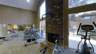 This fitness center combines functional design with distinctive architectural character. The centerpiece of the space is a dramatic floor-to-ceiling brick fireplace, adding a warm, rustic focal point that sets this gym apart from typical fitness facilities. Soaring vaulted ceilings amplify the sense of openness, while large sliding glass doors and tall windows flood the interior with abundant natural light and offer views of the surrounding outdoor greenery.

The workout area is well-appointed with a range of cardio equipment, including treadmills and stationary cycling bikes, arranged to make efficient use of the open floor plan. Mirrored walls along one side visually expand the space and provide useful feedback for those exercising. The neutral wall paneling and carpeted flooring contribute to a comfortable, inviting atmosphere.

The combination of the vaulted ceiling, expansive glazing, and lush outdoor views creates an airy environment that feels refreshing during a workout. The brick fireplace not only serves as an architectural statement but also adds a cozy ambiance to the space. Overall, this fitness center offers a thoughtfully designed environment that balances practical exercise amenities with warm, distinctive architectural details.