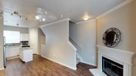 This two-story Dallas residence offers a well-organized layout spread across two levels, connected by a central staircase visible from the main living area. The ground floor showcases rich, dark hardwood-style flooring that runs throughout, providing a warm and cohesive aesthetic. Crown molding frames the ceilings, lending a polished, refined character to the space. The living area is anchored by a classic white-mantled fireplace, serving as a focal point and adding an inviting ambiance. A ceiling fan with integrated lighting fixture provides both airflow and illumination, complementing the recessed and flush-mount ceiling lights throughout. The neutral, light-gray wall palette creates a clean and versatile backdrop adaptable to a variety of interior styles. White baseboards and trim throughout provide sharp contrast and reinforce the home's crisp, classic detailing. A glimpse into an adjacent area near the staircase suggests additional functional space on the main level. The staircase, finished with white railings and risers, leads to the upper floor, where additional living quarters are situated. Overall, the space conveys a balanced mix of traditional architectural details and modern, move-in-ready finishes.