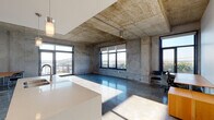 Located at 122 Delaware St in Kansas City's historic River Market district, this single-floor loft showcases a bold industrial aesthetic defined by exposed cast-concrete ceilings, raw concrete walls, and polished concrete floors throughout. The open-concept living and dining area is bathed in natural light from expansive black-framed windows that frame striking views of the Missouri River and a nearby bridge, creating a dramatic connection to the urban landscape. A sleek kitchen features white upper cabinetry, warm wood-toned lower drawers, stainless steel appliances including a range, microwave, and refrigerator, and a large white waterfall-edge island with an integrated sink. A dedicated workspace area benefits from the same panoramic views, offering an inspiring environment for productivity. The unit includes two full bathrooms: one featuring a soaking tub with subway tile surround and a wood-vanity with white countertop, and a second with a glass-enclosed walk-in shower, matching vanity, and polished chrome fixtures. Bedrooms continue the industrial theme with exposed concrete ceilings and large windows. Direct access to an outdoor terrace is also available. The overall ambiance is modern, open, and urban, with raw materials balanced by clean, contemporary finishes.