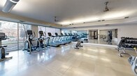 Located at 2986 W Lehman Ave in West Valley City, Utah, this dedicated fitness center offers a generously sized, single-floor exercise space designed to support a comprehensive workout experience. The room features polished large-format tile flooring that reflects the bright, well-distributed overhead fluorescent lighting, contributing to a clean and energizing atmosphere throughout.

A row of cardio equipment — including multiple treadmills and elliptical machines — lines the perimeter wall beneath expansive floor-to-ceiling windows, allowing natural light to flood the space and providing pleasant outdoor views of the surrounding neighborhood and landscaping. A wall-length mirror along one side visually expands the room and supports form-checking during workouts.

The strength training area is anchored by a multi-function cable and weight machine, complemented by a dumbbell rack with a wall-mounted flat-screen television nearby. A workout bench adds additional versatility for free-weight exercises. Ceiling fans are positioned throughout to ensure proper air circulation and comfort during use.

A textured ceiling adds a classic architectural touch, while glass-paneled interior doors and windows connect the fitness room to adjacent areas, maintaining an open and accessible feel. The space is thoughtfully organized to accommodate both cardio and strength training needs in a functional, community-oriented setting.