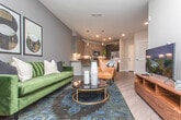 This single-floor residence combines contemporary design with functional living across a well-organized layout. The open-concept kitchen is a standout feature, anchored by a large central island with a white quartz countertop and seating for multiple guests. Flat-panel cabinetry in a warm wood-tone finish pairs with dark lower cabinets and stainless steel appliances, including a range and microwave. Pendant lighting above the island adds a refined touch, while recessed ceiling lights illuminate the entire space. The kitchen flows seamlessly into the living area, which features warm wood-look flooring, neutral wall tones, and a built-in shelving unit that doubles as a functional entryway organizer with hooks and woven storage baskets. The primary bedroom offers a striking design with a bold navy accent wall, natural light from large windows with floor-length curtains, wood-look flooring, and a ceiling fan. The entryway hallway is clean and efficient, featuring louvered closet doors that provide convenient in-unit storage. Throughout the residence, consistent wood-look flooring, crisp white trim, and a neutral color palette create a cohesive and modern aesthetic that is both inviting and practical.