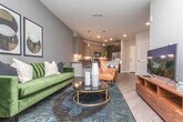 This single-floor residence combines contemporary design with functional living across a well-organized layout. The open-concept kitchen is a standout feature, anchored by a large central island with a white quartz countertop and seating for multiple guests. Flat-panel cabinetry in a warm wood-tone finish pairs with dark lower cabinets and stainless steel appliances, including a range and microwave. Pendant lighting above the island adds a refined touch, while recessed ceiling lights illuminate the entire space. The kitchen flows seamlessly into the living area, which features warm wood-look flooring, neutral wall tones, and a built-in shelving unit that doubles as a functional entryway organizer with hooks and woven storage baskets. The primary bedroom offers a striking design with a bold navy accent wall, natural light from large windows with floor-length curtains, wood-look flooring, and a ceiling fan. The entryway hallway is clean and efficient, featuring louvered closet doors that provide convenient in-unit storage. Throughout the residence, consistent wood-look flooring, crisp white trim, and a neutral color palette create a cohesive and modern aesthetic that is both inviting and practical.