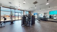 This contemporary fitness center in Hayward, California is thoughtfully designed to deliver a complete workout experience in a bright, welcoming environment. The single-floor layout is divided into distinct functional zones, each equipped with professional-grade Technogym machines and accessories. Large floor-to-ceiling windows along the street-facing wall flood the space with abundant natural light, offering views of the surrounding streetscape and creating an open, energizing atmosphere. The cardio zone features multiple treadmills and stair climbers positioned alongside the windows, complemented by a large wall-mounted display screen. A dedicated strength training area offers a comprehensive selection of cable machines, multi-use resistance stations, and a full dumbbell rack with benches. A separate enclosed room with glass-panel walls and hardwood-style flooring provides space for functional training, including stability equipment and additional machines. Mirrored walls throughout enhance the sense of depth and space, while pendant lighting fixtures lend a modern, polished aesthetic. Textured geometric accent walls in muted tones add visual interest alongside teal and neutral color accents. The herringbone-patterned rubber flooring offers durability and traction across the main workout areas. Built-in lockers and digital access panels round out the practical amenities of this well-equipped facility.