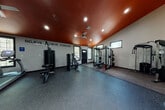 This dedicated fitness facility is thoughtfully designed to accommodate a full range of workout routines. The main training area features a striking burnt-orange ceiling accented by recessed lighting and a ceiling fan, creating an energetic atmosphere. Light-colored walls contrast boldly with the dark rubber flooring throughout, while the motivational phrase "Believe. Achieve. Conquer." spans one wall, reinforcing the space's purpose-driven character.

The primary workout zone is well-appointed with a power rack, cable machines, a weight bench, and a dumbbell rack offering a full range of free weights. A secondary area hosts cardio equipment, including an elliptical trainer, alongside a stability ball rack and medicine ball storage. Natural light enters through several windows, and a black entry door with decorative glass panels provides access from outside.

A transitional zone features double-height ceilings, a staircase, and dark accent walls adorned with decorative metallic wall art, adding an unexpected design element. Additional storage and utility doors are accessible from this area. The cohesive use of dark tones, industrial-style equipment, and purposeful layout gives the entire space a professional, motivating ambiance suited for serious training.