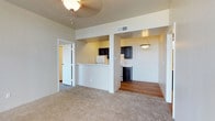 This single-floor residential unit offers a thoughtfully arranged open-concept layout that seamlessly connects the living area to the kitchen. The main living space features neutral-toned carpet underfoot and a ceiling fan overhead, creating a comfortable and functional environment. A half-wall pass-through connects the living area to the kitchen, enhancing the sense of openness while maintaining a defined separation between spaces. The kitchen is equipped with dark espresso-toned cabinetry that provides a striking contrast against the light walls, along with white appliances and a pendant light fixture that adds a warm, focused glow to the space. The kitchen and adjoining dining or utility area transition to rich hardwood-style flooring, adding warmth and visual depth. The walls throughout are finished in a clean, neutral palette that maximizes brightness and provides a versatile backdrop. A doorway visible from the main living area leads to an additional room or hallway, suggesting a practical and well-connected floor plan. Overhead HVAC venting is integrated discreetly into the ceiling. Overall, the unit presents a clean, move-in-ready aesthetic with a balance of soft textiles and hard surface finishes that lend the space both comfort and character.