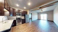 This single-floor space showcases a contemporary open-concept design that seamlessly blends the kitchen and living areas into one cohesive, flowing layout. The kitchen is appointed with rich, dark wood cabinetry that contrasts beautifully against stainless steel appliances, including a refrigerator, range, and over-the-range microwave. The warm hardwood-style flooring extends throughout the entire space, adding continuity and a sense of warmth.

An industrial touch is introduced through the exposed ventilation ductwork that runs along the ceiling, lending the space an urban loft character. Recessed lighting in the kitchen provides focused, practical illumination, while a ceiling fan in the living area ensures comfort and adds a casual, residential feel.

The living area is bright and open, benefiting from natural light that pours in through a sliding glass door, which also provides access to an outdoor area — ideal for extending the living space beyond the interior. A secondary doorway leads to an additional room, offering a sense of depth and privacy within the layout. The overall ambiance is modern and functional, combining industrial design elements with warm, inviting finishes to create a well-balanced and comfortable living environment.
