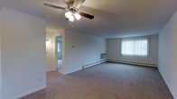 This single-floor unit offers a practical and functional layout designed for comfortable everyday living. The main living area is generously sized, featuring wall-to-wall carpeting that adds warmth underfoot. A ceiling fan with an integrated light fixture is centrally mounted, providing both air circulation and ambient lighting throughout the space. Natural light enters through a large window dressed with vertical blinds, creating a bright and airy atmosphere. Along the base of the exterior wall, baseboard heating units ensure consistent warmth, complemented by a wall-mounted air conditioning unit for year-round climate control. The walls are finished in a clean, neutral white palette, lending the space a simple and versatile backdrop. A hallway visible from the living area leads to additional rooms, with a wall-mounted light fixture illuminating the passage. A thermostat is conveniently positioned near the hallway entrance. The overall design is straightforward and efficient, prioritizing open, usable square footage with minimal architectural ornamentation, making it a functional and adaptable living space.