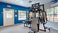 Located at 917 Country Club Park in DeLand, Florida, this single-story community clubhouse offers a well-appointed and versatile gathering space. The main lounge area is immediately welcoming, featuring soaring vaulted ceilings, large-format tile flooring, and an abundance of natural light. Ceiling fans are distributed throughout, enhancing comfort across the open layout. Comfortable seating arrangements, including sofas and accent chairs, create an inviting social atmosphere, while a wall-mounted television adds to the amenity offerings. French doors with decorative curtains lead to the exterior, further brightening the interior. A kitchenette area with white cabinetry, a refrigerator, and a raised bar with seating supports resident gatherings and events. Accessible through large glass-paneled French doors, the dedicated fitness room features bold blue accent walls, carpeted flooring, and a ceiling fan. It is equipped with a multi-station cable weight machine, a treadmill, an elliptical, and additional exercise equipment, alongside wall-mounted mirrors that amplify the sense of space. A wall-mounted television and seating area round out the fitness room. Together, these spaces form a functional and well-maintained community hub suited for both active and social use.