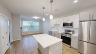 Located at 83 Pleasant Valley St in Methuen, Massachusetts, this well-designed one-bedroom, one-bathroom unit offers a thoughtful layout with modern finishes throughout. The kitchen serves as a central highlight, featuring sleek white cabinetry extending to the ceiling, a classic subway tile backsplash, quartz countertops, a kitchen island with pendant lighting, and a full suite of stainless steel appliances including a dishwasher, range, microwave, and refrigerator. Light hardwood-style flooring runs throughout the main living areas, enhancing the bright and open feel of the space. The living area flows naturally from the kitchen, creating an inviting, functional environment. The bedroom offers plush carpeting and a window with views of the surrounding greenery, allowing natural light to fill the room. A dedicated walk-in closet with rod storage provides ample organizational space. The bathroom is finished with a clean, minimalist aesthetic, featuring a wide vanity with a quartz countertop, an under-mount sink, warm cabinetry, a large mirror with overhead lighting, and polished chrome fixtures. Recessed lighting and neutral tones throughout lend the unit a cohesive, contemporary character.
