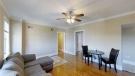 This single-story space presents a clean, understated interior anchored by warm hardwood flooring that extends throughout the main living area. The room is finished with classic crown molding along the ceiling perimeter, adding a refined architectural detail to the otherwise neutral, light gray walls. A five-blade ceiling fan with integrated lighting fixture serves as the central overhead light source, providing both illumination and airflow to the space.

The living area accommodates a gray sectional sofa paired with a patterned area rug, creating a casual and functional seating arrangement. A small, round black dining table and matching chair are positioned to one side, offering a compact dining or workspace solution. The layout is straightforward and efficient, making good use of the available floor space.

Two open doorways are visible from the main living area — one leading to a room with warm yellow-toned walls, and another partially open door suggesting an additional room or closet. White baseboards and door trim provide clean contrast against the hardwood floors. Ceiling vents are also present, indicating a ducted HVAC system. Overall, the space presents a modest, functional environment with traditional detailing.