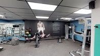 This fitness facility is a purpose-built workout space designed to inspire and support a comprehensive training regimen. The room features a striking floor-to-ceiling mural of Muhammad Ali, accompanied by his iconic motivational quote: "I hated every minute of training, but I said... Don't quit. Suffer now and live the rest of your life as a champion." This bold graphic centerpiece sets a determined, athletic tone throughout the space.

The facility is outfitted with a well-rounded selection of equipment to support both strength and cardio training. A multi-station cable and weight machine is positioned along one side, offering versatile resistance training options. A rowing or cycling machine provides cardiovascular workout capability, while a rack of medicine balls in varying weights supports functional fitness and conditioning exercises.

The flooring consists of durable rubber gym matting, ideal for high-impact activity and equipment use. Overhead, a drop ceiling with large panel lighting ensures the space is evenly and brightly illuminated. The walls are painted in a deep charcoal and blue color scheme, contributing to a focused, energetic atmosphere. Mirror panels along one wall enhance the sense of space and allow users to monitor their form during workouts. The single-floor layout ensures all amenities are conveniently accessible in one open area.