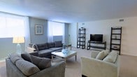 This single-floor space offers a warm and inviting atmosphere defined by a cohesive neutral color palette and thoughtful layout. The living room serves as the centerpiece of the home, featuring soft blue-gray walls that complement the light-toned carpeted flooring, creating a calm and relaxing ambiance. A large window dressed with sheer white curtains allows natural light to fill the room, enhancing the open and airy feel of the space. The living area is comfortably furnished with a generously sized gray sectional sofa and a matching accent chair, arranged around a mirrored glass coffee table that adds a touch of elegance. A ladder-style bookshelf provides both storage and decorative display space, while a dedicated media area with a flat-screen television and a sleek media stand rounds out the room's functionality. Subtle coastal-inspired wall art contributes to the overall relaxed aesthetic of the space. The layout is practical and well-organized, making efficient use of the available space while maintaining an uncluttered, comfortable environment suitable for everyday living and entertaining.