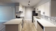 This single-floor unit offers a thoughtfully designed layout that balances comfort and functionality. The open-concept living area features durable wood-look flooring, neutral gray walls, a textured ceiling, and a ceiling fan with integrated lighting. Vertical blinds along a sliding glass door allow natural light to fill the room while providing access to the outdoors.

The kitchen is a standout feature, showcasing white shaker-style cabinetry with matte black hardware, crisp white countertops, and an eye-catching patterned tile backsplash. Stainless steel appliances — including a refrigerator, dishwasher, and range — are complemented by track lighting overhead. A peninsula counter provides additional prep space and separates the kitchen from the living area.

The bedroom continues the neutral palette with soft carpeting, a ceiling fan, and a window fitted with vertical blinds for natural light and privacy. The bathroom is well-equipped with a full tub and shower combination surrounded by large-format gray tile, while a separate vanity area offers a white cabinetry unit, a single sink with white countertop, and a lighted mirror — all finished with wood-look flooring for a cohesive aesthetic throughout the unit.