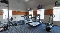 This fitness center is designed to provide a functional and motivating environment for exercise. The space features a clean, modern aesthetic with walls painted in a two-tone scheme of warm orange and neutral gray, creating an energetic yet focused atmosphere. The flooring consists of durable striped carpet in shades of blue and gray, well-suited for a high-traffic fitness environment.

The cardio area is equipped with multiple treadmills aligned to face a row of windows fitted with white horizontal blinds, allowing natural light to fill the room while maintaining privacy. Ceiling-mounted fans provide additional ventilation and comfort during workouts. Wall-mounted flat-screen televisions are positioned at strategic angles throughout the space, ensuring visibility from various workout stations and enhancing the overall exercise experience.

Additional fitness equipment, including free weights and a cable resistance machine, is available along the sides of the room, offering users a range of strength and conditioning options. The layout is thoughtfully organized to maximize usable floor space while maintaining safe distances between equipment. Overall, the fitness center offers a practical and well-appointed setting for cardio and strength training activities.