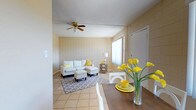 This single-floor space offers a comfortable and cohesive layout with an open-concept living and dining area that flows naturally together. The living area is anchored by light beige tile flooring and a textured ceiling, with a ceiling fan providing both functionality and a classic touch. Large windows allow natural light to fill the room, enhancing the warm, neutral color palette of the walls and décor. The living space features a white sectional sofa accented with yellow and white throw pillows, paired with a matching ottoman and a patterned area rug that adds visual interest to the room. A side table with a table lamp completes the seating arrangement, creating an inviting and relaxed atmosphere. Adjacent to the living area, the dining space is defined by a dark wood table paired with white chairs, adding a pleasant contrast to the light surroundings. A vibrant arrangement of yellow calla lilies serves as a centerpiece, echoing the cheerful yellow accents found throughout the space. The walls feature a subtle tile or panel texture that adds architectural character without overwhelming the overall aesthetic. The space presents a clean, well-organized interior that balances functionality with a bright, welcoming ambiance.
