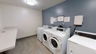 This shared laundry room is a functional and well-maintained facility designed to meet residents' everyday laundry needs. The room features a clean, modern aesthetic with a two-tone color scheme — light upper walls paired with a muted steel-blue lower accent wall — creating a tidy and pleasant environment. Durable wood-look flooring runs throughout, offering both practicality and a contemporary touch.

The room is equipped with multiple front-loading washing machines and dryers, each outfitted with digital control panels for ease of use. The machines are arranged side by side along the main wall, providing an efficient layout that maximizes the available space. Informational notices and usage guidelines are posted on the accent wall above the machines, ensuring residents have clear instructions at hand.

Overhead lighting provides bright, even illumination throughout the space, making it comfortable to use at any time of day. The overall layout is straightforward and accessible, with an open floor plan that allows for easy movement while loading and unloading laundry. The room's clean design and well-organized setup reflect a commitment to providing a convenient and user-friendly shared amenity.