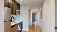 Located at 1831-1835 Harmon St in Berkeley, CA, this thoughtfully designed single-floor space centers around a long, light-filled hallway that serves as the connective spine of the layout. The corridor is finished with light wood luxury vinyl plank flooring that runs its full length, complemented by crisp white walls, clean white baseboards, and recessed ceiling lighting that provides consistent, even illumination throughout. The hallway's elongated form creates a sense of depth and openness, and framed artwork adorns the walls, adding character and visual interest to the passage. Stylish slate-blue panel doors punctuate the corridor, providing access to the adjoining rooms and contributing a subtle contrast to the otherwise neutral palette. The space includes two full bathrooms, one of which features a tub and shower combination enclosed by sleek sliding glass doors with matte black hardware, white tile surround, geometric mosaic floor tile, and open wooden shelving with brass accents against soft blue walls. A glimpse of a kitchenette with dark cabinetry, stainless steel appliances, and a light backsplash is visible adjacent to the hallway, rounding out this efficient and modern Berkeley property.