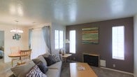 This single-floor space offers a warm and inviting atmosphere centered around a well-appointed living room. The living area serves as the heart of the home, featuring a classic fireplace with a wood mantel that anchors the room and provides a focal point for relaxation. A textured ceiling adds visual interest overhead, while multiple large windows — dressed with light blue curtains and vertical blinds — allow generous amounts of natural light to fill the space, creating a bright and airy feel. The living room is furnished with a plush gray sofa accented with decorative throw pillows, complemented by a wooden coffee table and a side table with a classic lamp. A bold, colorful abstract canvas painting adorns the taupe accent wall, adding a contemporary artistic touch. Neutral-toned carpeting runs throughout, contributing to the space's comfortable and cohesive aesthetic. The layout is thoughtfully arranged to maximize both functionality and comfort, making it well-suited for everyday living and casual entertaining. Overall, the space balances a neutral color palette with warm accents, creating an environment that feels both stylish and approachable.