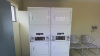 This single-floor laundry facility is designed with practicality and convenience in mind. The space is equipped with two sets of stacked, coin-operated washer and dryer units, providing ample capacity for laundry tasks. The machines are commercial-grade, stacked configurations that maximize the use of vertical space, keeping the floor area open and easy to navigate. The units feature coin-slot mechanisms, making them well-suited for shared or communal use. The room's neutral-toned walls and utilitarian finishes contribute to a clean, no-frills environment focused on function. A metal chair is available along the side wall, offering a place to sit while waiting for laundry cycles to complete. The flooring appears durable and easy to maintain, consistent with the practical nature of the space. Overall, this laundry facility provides a straightforward, efficient, and well-equipped area to handle everyday laundry needs.