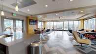 This single-floor communal space is designed with a lively, contemporary aesthetic that balances energetic design elements with functional versatility. The most immediately striking feature is the large-scale chevron-patterned flooring, rendered in contrasting light and dark tones, which anchors the entire space and gives it a strong visual identity throughout.

The lounge area is organized into distinct zones. A vibrant seating section features a bold orange sofa accented with teal and patterned throw pillows, positioned beneath a wall-mounted flat-screen TV set against a rich yellow accent wall. Textured orange drapery panels frame the expansive floor-to-ceiling windows, which flood the interior with natural light and offer sweeping views of an urban skyline. Additional seating includes modern swivel chairs, a sleek white lounge chair, and a game table set with a chess board, creating an inviting social environment.

On the opposite end, a kitchenette area provides practical amenities, featuring dark cabinetry, a stainless steel refrigerator, a bold red subway tile backsplash, and a counter with bar seating. Recessed ceiling lighting throughout ensures consistent illumination, complementing the abundant natural light. The overall ambiance is dynamic, colorful, and thoughtfully arranged for both casual interaction and relaxed downtime.