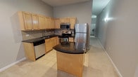 This single-floor residence offers a thoughtfully arranged layout that connects the kitchen, bedroom, and bathroom in a functional and efficient flow. The kitchen serves as a standout feature, showcasing light maple cabinetry that extends to the ceiling for generous storage capacity. Dark granite countertops provide a striking contrast against the cabinetry, while a full suite of stainless steel appliances — including a refrigerator, electric range, built-in microwave, and dishwasher — ensures a well-equipped cooking environment. A curved kitchen island with matching granite adds extra prep space and visual interest. Tile-patterned backsplash and light tile flooring complete the kitchen's clean, polished aesthetic. The bathroom is finished with beige ceramic wall tiles accented by a navy blue tile border in the tub surround, complemented by tile flooring, a full soaking tub with shower, and dark granite vanity countertop details. The bedroom offers plush carpeting, a ceiling fan, a walk-in closet, and additional built-in storage, providing a comfortable and organized retreat. Neutral gray walls and light carpet throughout create a cohesive, move-in-ready atmosphere across the entire space.