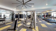 This fitness center offers a spacious, single-floor layout designed to accommodate a full range of workout needs. The interior features a bold, geometric carpet in contrasting shades of yellow, gray, and blue, adding a dynamic visual element to the space. Large ceiling fans with industrial-style blades are mounted overhead, promoting air circulation throughout the room and contributing to a comfortable workout environment.

The strength training area is well-appointed with cable machines, a functional trainer, adjustable benches, and a variety of free weights and accessories. Mirrored walls line sections of the space, enhancing the sense of openness and allowing users to monitor their form during exercise. The walls are finished in a two-tone design with dark navy and light gray tones, giving the facility a polished, modern aesthetic.

Toward the back of the space, cardio equipment including treadmills and additional machines can be seen, suggesting a well-rounded offering for both strength and endurance training. Recessed lighting complements the overhead fans, ensuring the space is well-lit throughout. The overall ambiance is clean, modern, and professional, reflecting a thoughtfully designed fitness facility.