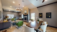This single-story space is defined by its impressive vaulted ceilings with dark exposed beams, creating a sense of volume and grandeur throughout. The open-concept layout seamlessly connects the living and dining areas, all finished with large-format travertine-style tile flooring that adds an elegant, cohesive feel underfoot.

The living area is anchored by a striking fireplace with a dark-painted mantel and granite surround, flanked by windows dressed with white blinds that invite natural light. A round mirror above the mantel adds a decorative focal point. Seating includes bold green leather accent chairs, a grey sofa with yellow and patterned throw pillows, and plaid armchairs, creating an eclectic yet refined atmosphere. Ceiling fans and a warm wooden chandelier with glass accents provide both comfort and stylish illumination.

The dining area features a substantial wood table surrounded by white upholstered chairs, lit by a matching chandelier overhead. Arched doorways and structural columns define separate zones while maintaining visual openness. Neutral grey walls provide a versatile backdrop for curated artwork, botanical prints, and greenery, lending the space a welcoming, well-appointed ambiance throughout.