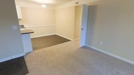 This single-floor residential space offers a clean, functional layout designed for comfortable everyday living. The main living and dining area is finished in warm, neutral tones with cream-colored walls accented by white chair rail molding, creating a classic and tidy aesthetic. The flooring transitions from plush, beige carpet in the living area to a darker hard-surface flooring in the dining zone, clearly delineating the two spaces while maintaining a cohesive visual flow. A ceiling-mounted light fixture provides overhead illumination to the dining area, ensuring the space remains bright and well-lit. White baseboards trim the perimeter of the room, adding a polished finishing touch throughout. A doorway along the far wall opens to an adjacent room or hallway, hinting at additional space beyond the main living area. A thermostat or control panel is visible on the wall near the doorway, reflecting practical built-in amenities. The countertop edge visible at the edge of the frame suggests a nearby kitchen or bar area, adding to the overall functionality of the layout. The space presents itself in a well-maintained, move-in-ready condition with a straightforward, livable design.