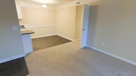 This single-floor residential space offers a clean, functional layout designed for comfortable everyday living. The main living and dining area is finished in warm, neutral tones with cream-colored walls accented by white chair rail molding, creating a classic and tidy aesthetic. The flooring transitions from plush, beige carpet in the living area to a darker hard-surface flooring in the dining zone, clearly delineating the two spaces while maintaining a cohesive visual flow. A ceiling-mounted light fixture provides overhead illumination to the dining area, ensuring the space remains bright and well-lit. White baseboards trim the perimeter of the room, adding a polished finishing touch throughout. A doorway along the far wall opens to an adjacent room or hallway, hinting at additional space beyond the main living area. A thermostat or control panel is visible on the wall near the doorway, reflecting practical built-in amenities. The countertop edge visible at the edge of the frame suggests a nearby kitchen or bar area, adding to the overall functionality of the layout. The space presents itself in a well-maintained, move-in-ready condition with a straightforward, livable design.