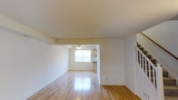 This two-story space is designed with a straightforward, functional layout that emphasizes openness and ease of movement. The main living area is characterized by polished hardwood floors that reflect natural light, lending warmth and brightness to the room. Neutral white walls throughout create a clean, versatile backdrop suitable for a variety of interior styles.

A ceiling fan is centrally positioned in the living area, providing both ventilation and a subtle decorative element. The open-plan design allows the living space to flow naturally toward the kitchen area, which is partially visible through a structural opening in the dividing wall. The kitchen features cabinetry and receives natural light from a window, keeping the area feeling bright and airy.

A staircase is visible along one side of the living area, providing access to the second floor and contributing to the vertical dimension of the space. The overall ambiance is light, neutral, and move-in ready, with hardwood flooring and consistent wall finishes tying the layout together cohesively across the main level.