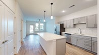 This single-floor unit showcases a thoughtfully designed open-concept layout that seamlessly connects the kitchen and living area. The kitchen is a standout feature, equipped with gray shaker-style cabinetry extending to the upper walls, white quartz countertops, a white subway tile backsplash, and a full suite of stainless steel appliances including a refrigerator with ice maker, dishwasher, and range. A generously sized central island with a bright white surface serves as both a prep space and gathering point, illuminated by two sleek pendant lights overhead.

The living area flows naturally from the kitchen and is finished with wood-look flooring, a ceiling fan, and large windows and a glass door that invite abundant natural light and outdoor views. Bedrooms are carpeted for comfort, with one featuring a ceiling fan and a bank of windows overlooking the exterior. A dedicated walk-in closet with wire shelving provides ample storage. The bathroom includes a soaking tub with subway tile surround, a gray vanity with quartz countertop, wood-look flooring, and direct access to the walk-in closet. Throughout, the unit maintains a clean, neutral palette that creates a bright and cohesive atmosphere.