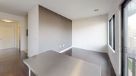 Dwell Studio is a thoughtfully designed single-floor space that blends modern functionality with clean, minimalist aesthetics. The kitchen is a standout feature, outfitted with stainless steel appliances including an electric range with overhead hood vent, a dishwasher, and a sink. Open stainless steel wall shelving provides practical storage, while a large stainless steel prep table anchors the center of the room. Dark hardwood flooring runs throughout, and a full-height sliding glass door opens to a private balcony, flooding the space with natural light and offering outdoor views. Adjacent to the kitchen, the open living area is defined by a distinctive accent wall in a warm taupe tone, contrasting with the bright white walls throughout the rest of the unit. Dark hardwood flooring continues seamlessly into this space, creating a cohesive flow. The bedroom features plush carpeting, a built-in closet with bifold doors, and an electrical panel mounted on the wall. The full bathroom is clean and utilitarian, with a walk-in shower, a vanity with a wood-toned cabinet, and neutral tile flooring. Overall, the space maintains a bright, modern character with a practical layout suited for everyday living.