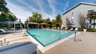 This inviting outdoor amenity space in Columbia, Maryland offers a thoughtfully designed retreat centered around a generously sized swimming pool with crystal-clear turquoise water. The pool deck is finished with a smooth concrete surface and is lined with rows of modern white lounge chairs, providing ample space for sunbathing and relaxation. A covered cabana with seating and shade umbrellas anchors one end of the deck, offering a comfortable respite from the sun. Additional lounge chairs with umbrellas are scattered throughout the patio area, ensuring plenty of shaded options for guests. The surrounding landscape is lush and well-maintained, with mature trees, flowering shrubs, and colorful potted plants adding vibrancy and a sense of privacy to the environment. Stone-accented architectural details on the adjacent building complement the natural greenery and elevate the overall aesthetic. A fitness facility is accessible directly from the pool area, enclosed by large floor-to-ceiling glass windows framed in dark metal, allowing natural light to fill the interior. Landscaped walkways with efficient drainage channels connect the various areas of the space, creating a cohesive and functional outdoor environment. The two-level building adjacent to the pool further enhances the sense of scale and community within this well-rounded amenity space.