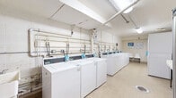 Located at 1189 Washington St in Middletown, Connecticut, this shared laundry facility offers a practical and well-equipped space designed to serve multiple users efficiently. The room is laid out in a straightforward rectangular floor plan with durable speckled tile flooring throughout, providing easy maintenance and a clean appearance.

A row of top-load washing machines lines one wall, each equipped with digital card-operated payment panels, offering a convenient cashless payment option. Opposite the washers, a bank of stacked dryers provides ample drying capacity. Exposed utility piping runs along the walls, connecting the machines to the building's plumbing and ventilation systems. A deep utility sink with a faucet is positioned along one wall, useful for pre-treating garments or hand-washing delicate items.

A white folding table with hairpin legs offers a dedicated surface for sorting and folding laundry, while a pair of white seating chairs provides a comfortable waiting area. Informational signage is posted on the walls, including instructions for using a laundry app for added convenience.

Two vending machines — one offering snacks and another stocked with beverages — add an extra layer of convenience for users during their visit. Overhead lighting illuminates the space evenly, and a security camera ensures a safe environment. The facility is functional, clean, and thoughtfully equipped for shared residential use.