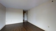 This single-floor space presents a straightforward and functional layout with a clean, understated aesthetic. The main living area is finished with dark hardwood-style flooring that contrasts with the bright white ceiling and neutral, off-white walls, creating a simple yet warm atmosphere. The open wall surfaces provide ample flexibility for furniture arrangement and personalization.

A doorway visible toward the rear of the space leads to an adjacent area, suggesting a connected hallway or secondary room. The transition zone between the two areas features lighter tile or vinyl flooring, offering a practical surface change that distinguishes the spaces. White baseboards run along the perimeter of the room, adding a clean finishing detail.

The space is well-lit with a bright, even light that highlights the openness of the room. The smooth white ceiling and minimal architectural detailing give the interior a modern, no-frills character. Electrical outlets and light switch panels are positioned at standard heights along the walls, reflecting a practical and functional design. Overall, the space offers a blank canvas well-suited for customization.
