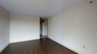 This single-floor space presents a straightforward and functional layout with a clean, understated aesthetic. The main living area is finished with dark hardwood-style flooring that contrasts with the bright white ceiling and neutral, off-white walls, creating a simple yet warm atmosphere. The open wall surfaces provide ample flexibility for furniture arrangement and personalization.

A doorway visible toward the rear of the space leads to an adjacent area, suggesting a connected hallway or secondary room. The transition zone between the two areas features lighter tile or vinyl flooring, offering a practical surface change that distinguishes the spaces. White baseboards run along the perimeter of the room, adding a clean finishing detail.

The space is well-lit with a bright, even light that highlights the openness of the room. The smooth white ceiling and minimal architectural detailing give the interior a modern, no-frills character. Electrical outlets and light switch panels are positioned at standard heights along the walls, reflecting a practical and functional design. Overall, the space offers a blank canvas well-suited for customization.