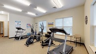 This fitness room is a dedicated single-level exercise space designed to support a range of cardio-focused workouts. The room is finished with neutral, cream-colored walls and durable speckled flooring that gives the space a clean, practical aesthetic suited for regular use. Fluorescent strip lighting is evenly distributed across the ceiling, ensuring the entire room is well-lit at all hours.

The equipment layout is thoughtfully arranged to maximize the available floor space. Key cardio machines include treadmills, elliptical trainers, and a stationary cycle, all positioned to allow comfortable use without crowding. A large wall-spanning mirror on one side of the room amplifies the sense of space and allows users to monitor their form during exercise. Two flat-screen TVs are mounted on the walls, adding an entertainment option for those working out.

Multiple windows fitted with blinds line the perimeter, allowing natural light to filter into the room while maintaining privacy. A water dispenser is conveniently placed near the entrance, and coat hooks on the wall provide practical storage for personal items. Informational fitness posters are displayed throughout the room. A wood-framed door with glass panels serves as the entry point, contributing a subtle warmth to the otherwise functional interior.