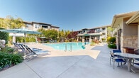 Located at 5102 Dowdell Ave in Rohnert Park, California, this two-story residential community showcases a thoughtfully designed outdoor amenity space that serves as its centerpiece. The community's architecture blends stucco exteriors with warm earth tones, accented by teal and wood-toned trim details, creating a cohesive California contemporary aesthetic across multiple interconnected buildings.

The outdoor area is anchored by a generously sized, freeform swimming pool with sparkling blue water, complemented by an adjacent spa. The expansive pool deck provides ample room for lounging, outfitted with blue chaise lounge chairs, shaded patio umbrellas, and bistro-style seating areas. A covered cabana with a ceiling fan and decorative lattice detailing offers a shaded retreat, supported by stone-clad columns and framed by vibrant plantings.

Lush, well-maintained landscaping is a defining feature throughout the grounds, with mature trees including palms, ornamental grasses, colorful flowering plants, and hedgerows creating a private, resort-like atmosphere. Wrought iron gated entries add a layer of security while maintaining visual appeal. A dedicated pool house facility serves the amenity area. Under Northern California's abundant sunshine, the entire outdoor space exudes a relaxed, welcoming ambiance ideal for year-round enjoyment.