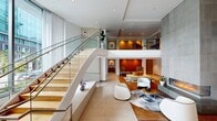 Located at 399 Fremont Street in San Francisco's SoMa district, this 2,340-square-foot residential lobby showcases sophisticated contemporary architecture across a single, thoughtfully designed floor. The space is anchored by a dramatic double-height atrium flooded with natural light through floor-to-ceiling windows that frame views of the surrounding cityscape. A large-format, stone-clad linear fireplace serves as a striking focal point in the central lounge area, which is furnished with sculptural white curved seating, a cowhide area rug, and a warm wood-toned floor that adds visual contrast to the otherwise bright, neutral palette. Curated artwork is displayed throughout, including a bold red floral canvas and a delicate butterfly wall installation that ascends toward the upper mezzanine level, accessible via a glass-railed staircase. Large-format tile flooring with dark inlay detailing extends through the main entry corridor, which features stepped transitions connecting different sections of the lobby. A glass-enclosed concierge station offers residents a dedicated service area with ample natural light. An additional service alcove provides residents with access to mailboxes and building amenities. Recessed lighting throughout enhances the clean, modern aesthetic, creating a welcoming and polished atmosphere.
